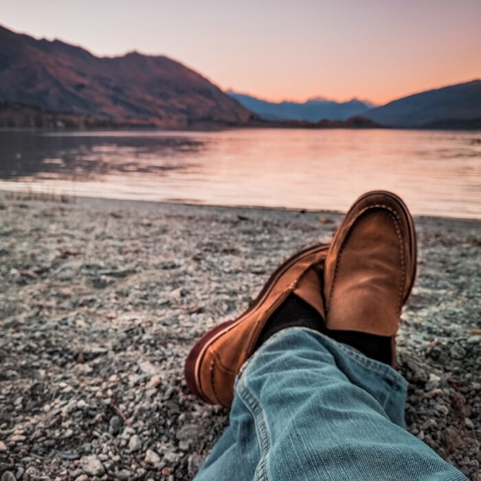 person wearing brown loafers sitting beside body of water